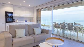 A modern living room with a sofa, a small round table, and a balcony with a view of the sea.