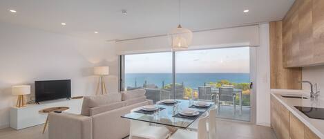 A modern living room with a glass dining table, a sofa, a TV, and a view of the ocean.