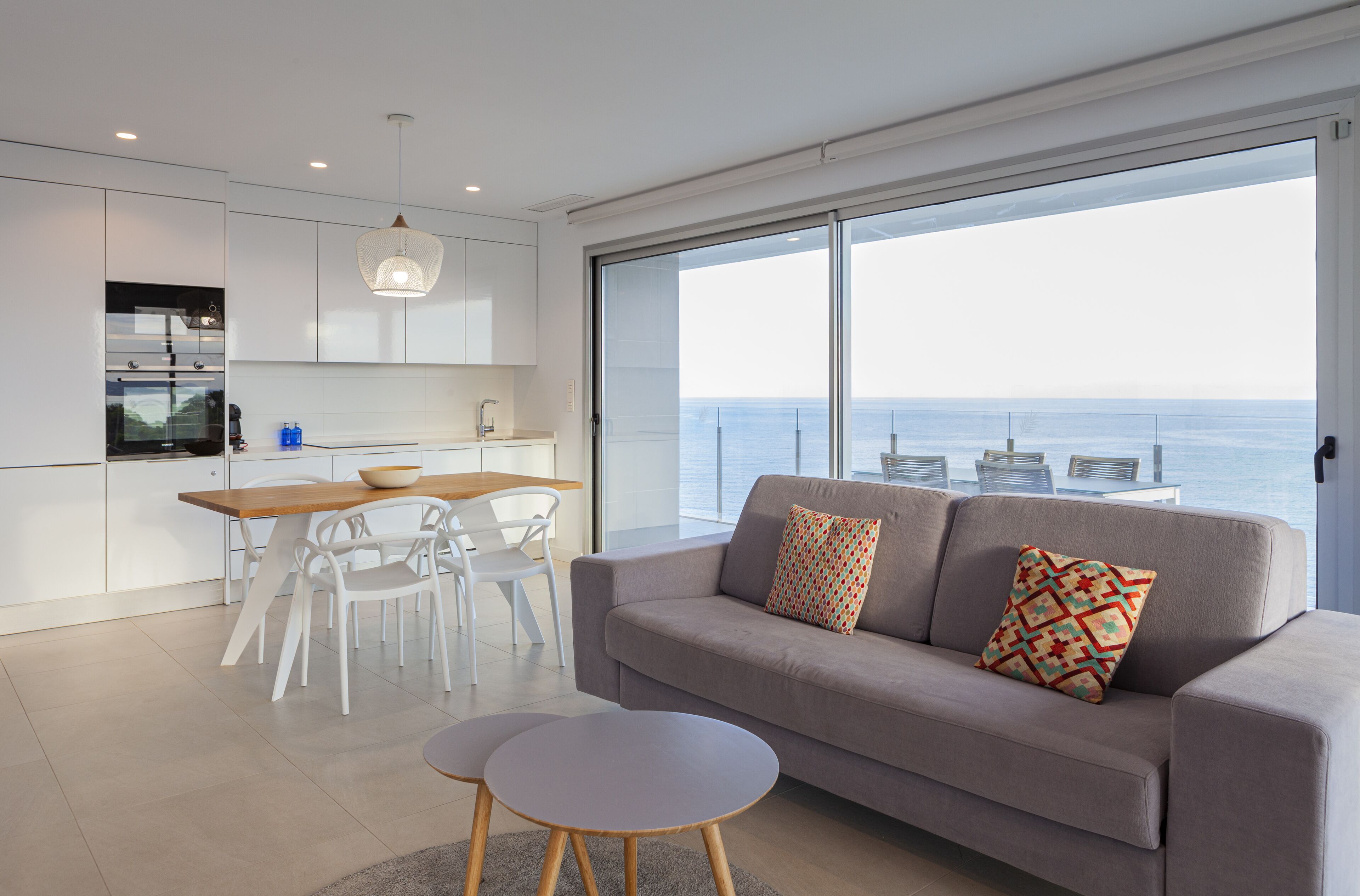 Una cocina moderna con armarios blancos, un comedor con mesa y sillas de madera, y un sofá gris con cojines estampados.
