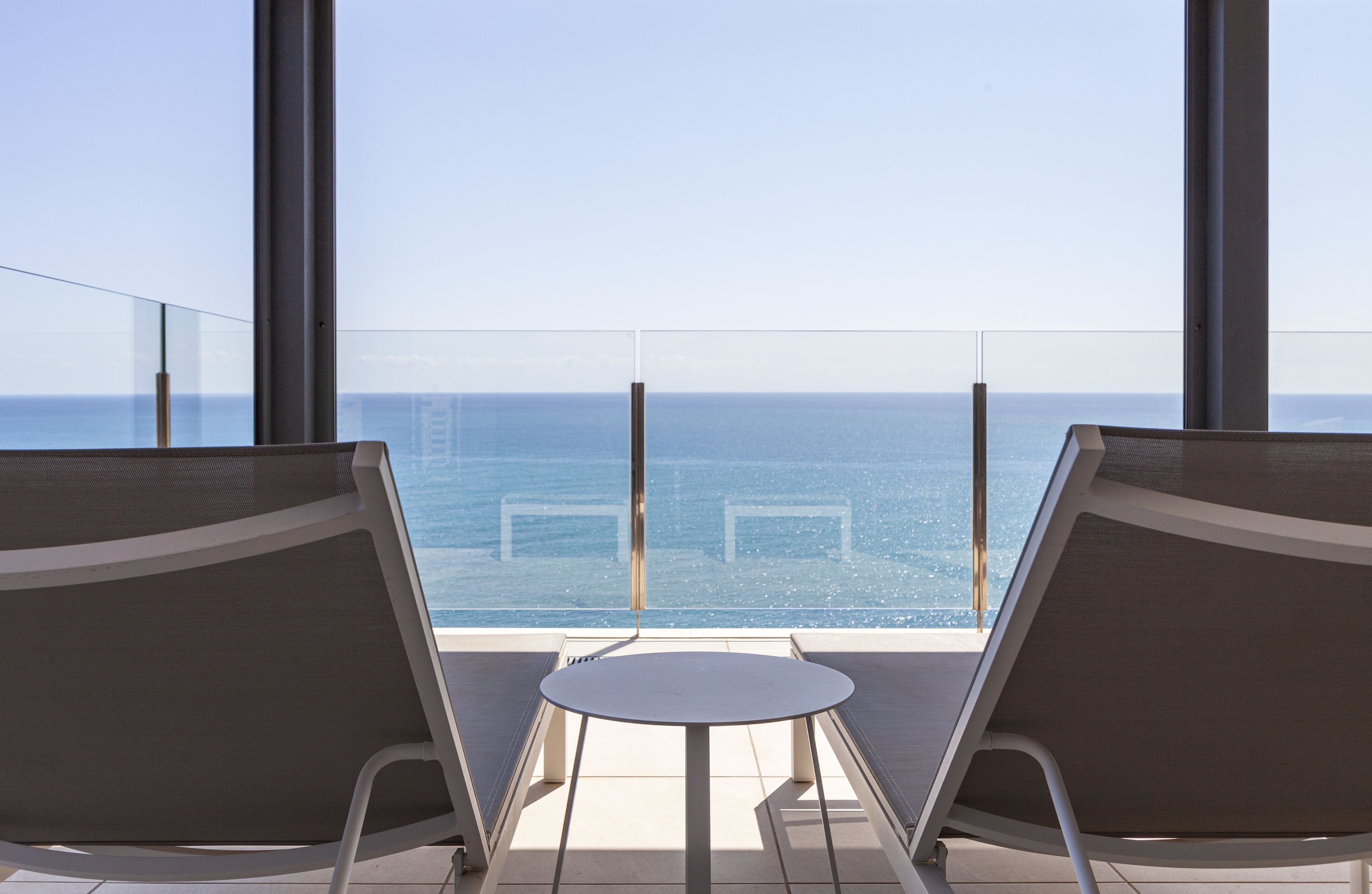 Un balcón con dos sillones de descanso y una mesita, con vistas al mar azul y edificios a lo lejos.