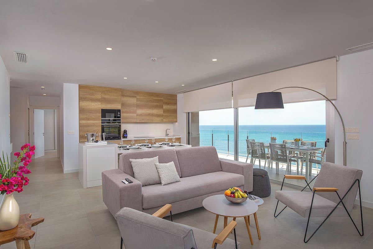 Ein modernes Wohnzimmer mit Sofa, Sessel und rundem Tisch, einer Küchenzeile sowie einem Balkon mit Meerblick.