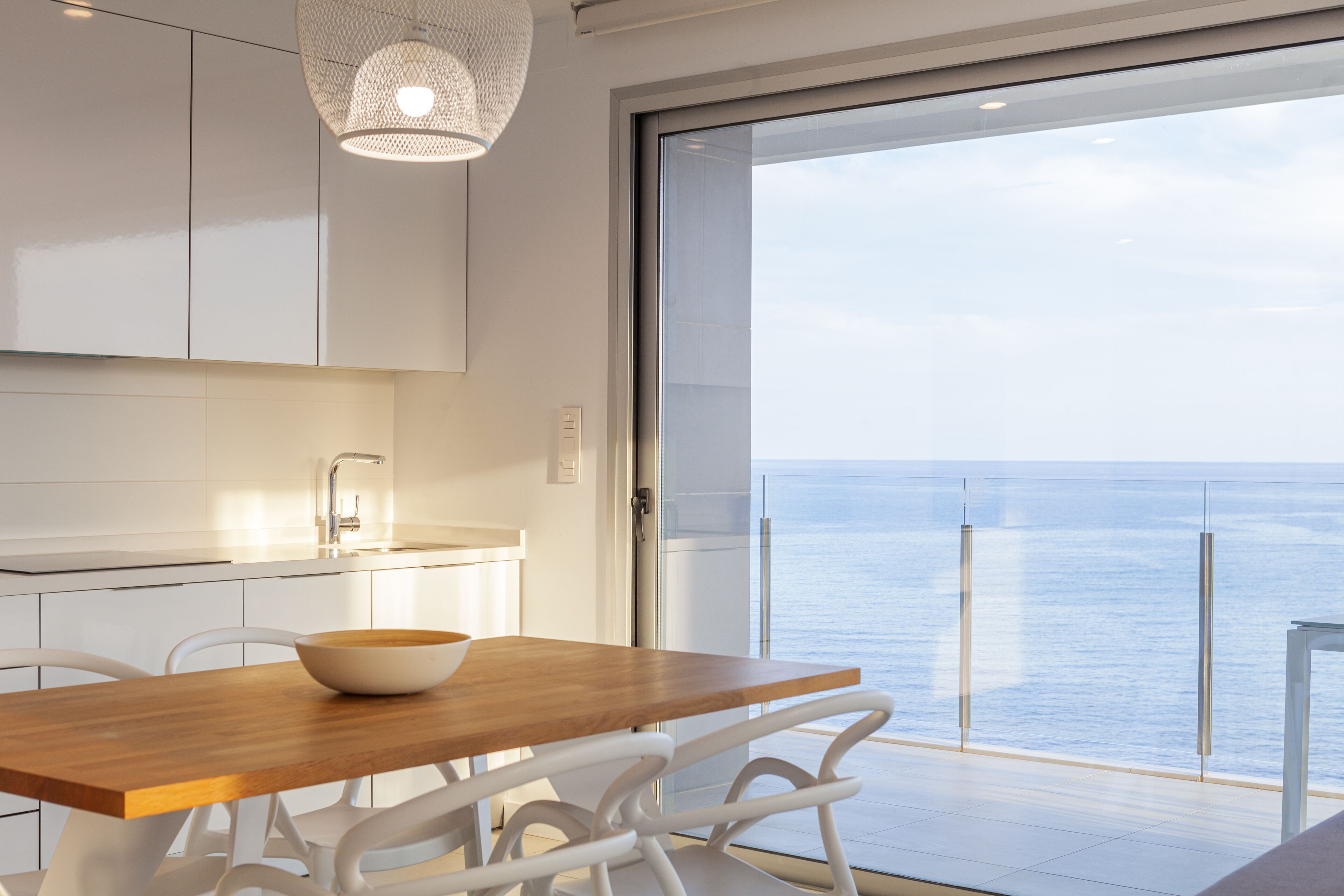 Une cuisine moderne avec des meubles blancs, une table en bois et un saladier. Une grande porte vitrée donne accès à un balcon avec vue sur la mer.