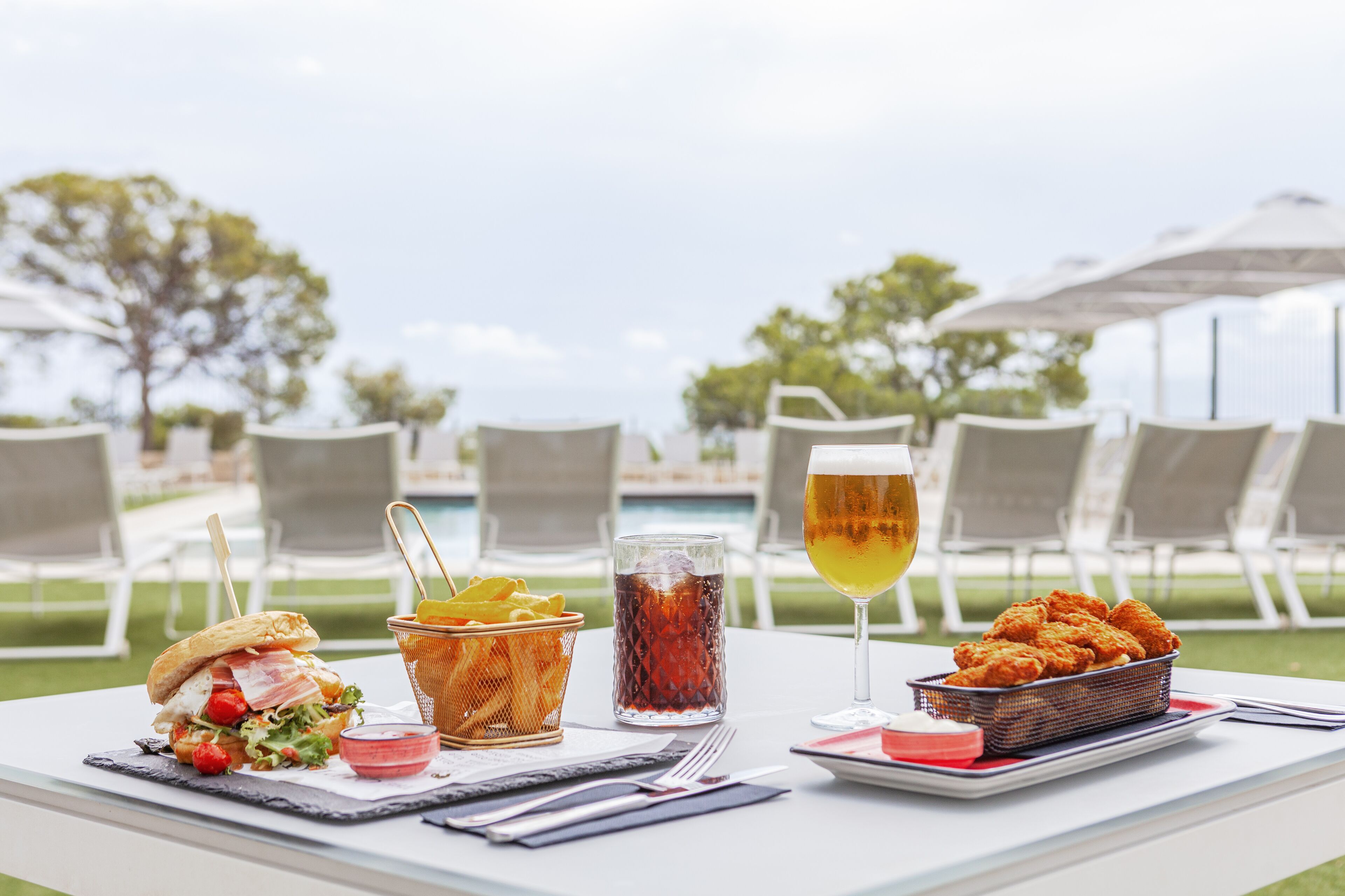 Een tafel gedekt met een sandwich, een mandje friet, een glas ijsthee en een glas bier, met buiten zitplaatsen en parasols op de achtergrond.