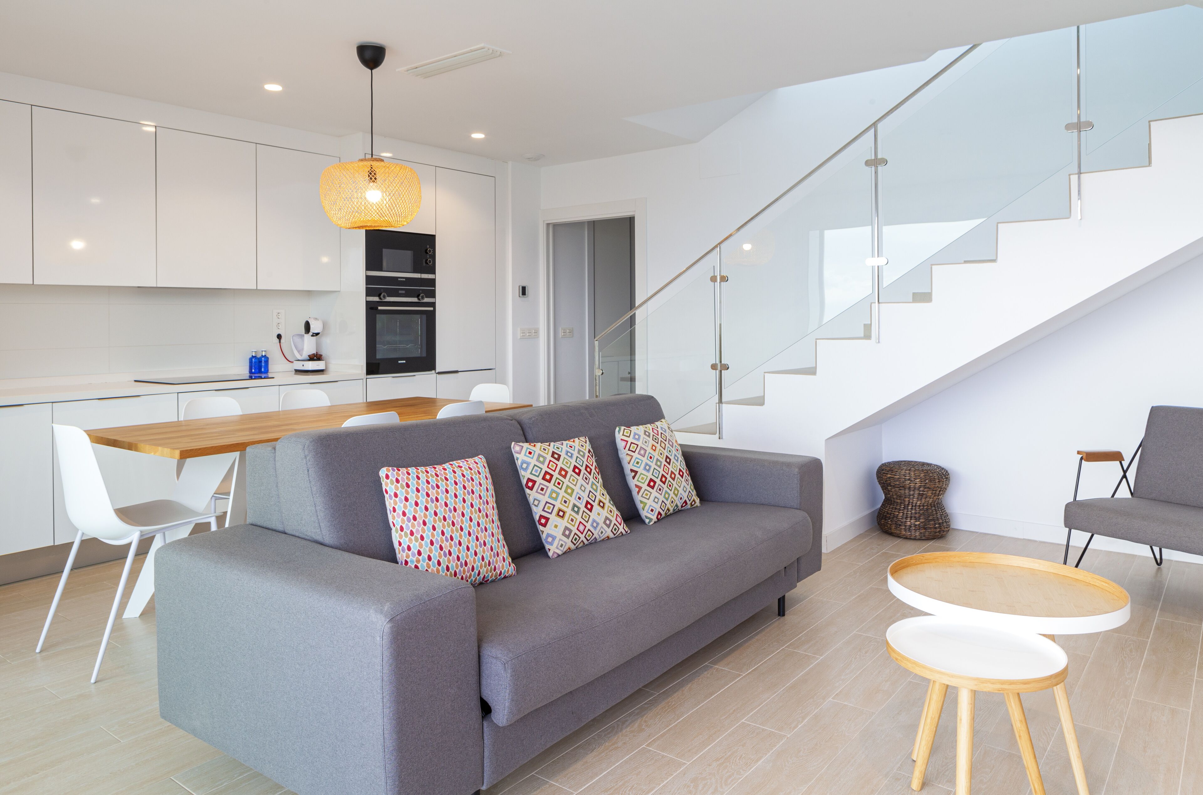 A modern living room with a grey sofa, a wooden dining table, and a staircase leading to an upper floor.