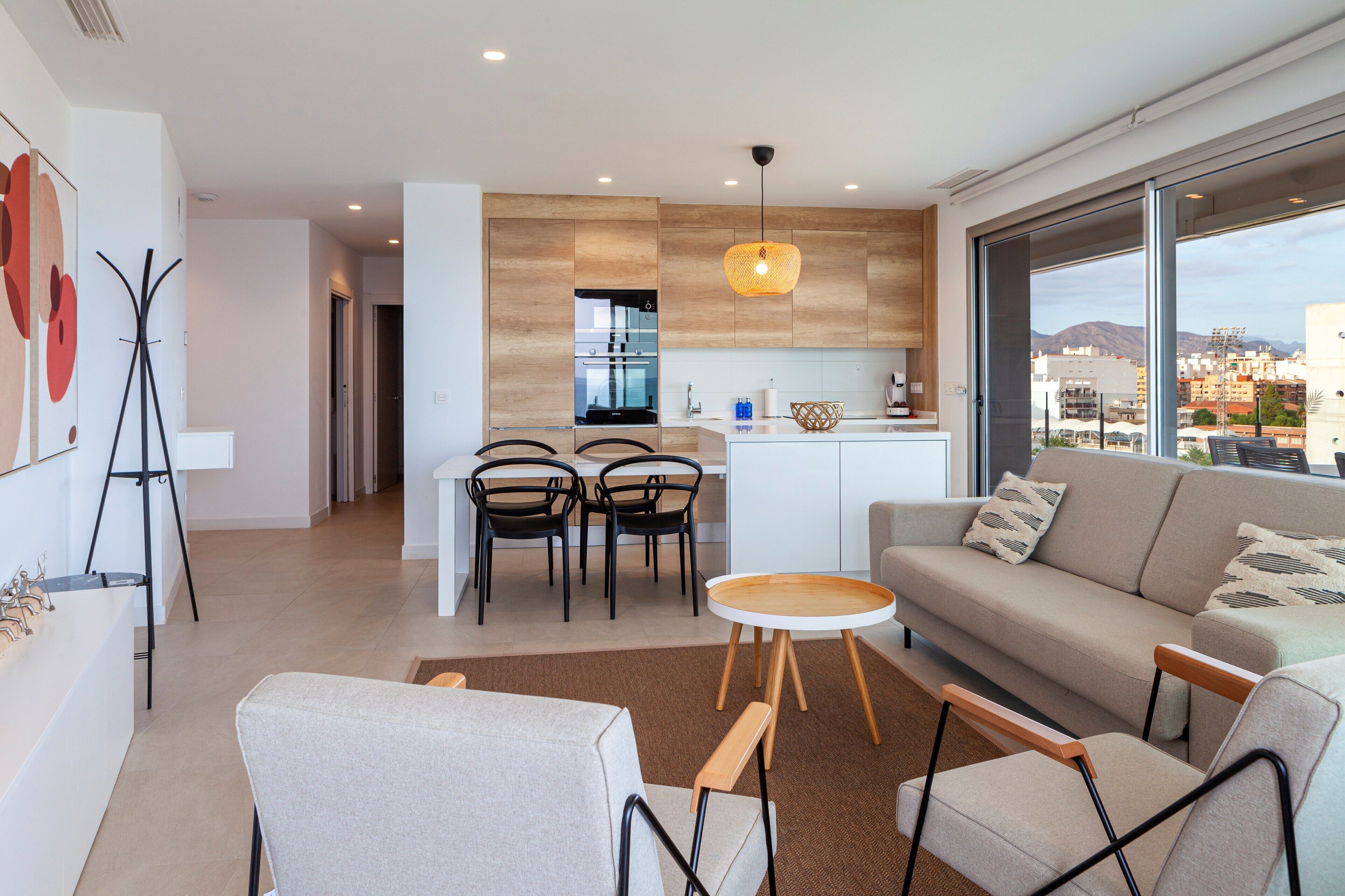 Un salón moderno con sofá, mesa de centro y sillas. Hay una zona de cocina con mesa y sillas para comer. La habitación tiene ventanas amplias con vistas a edificios y montañas.