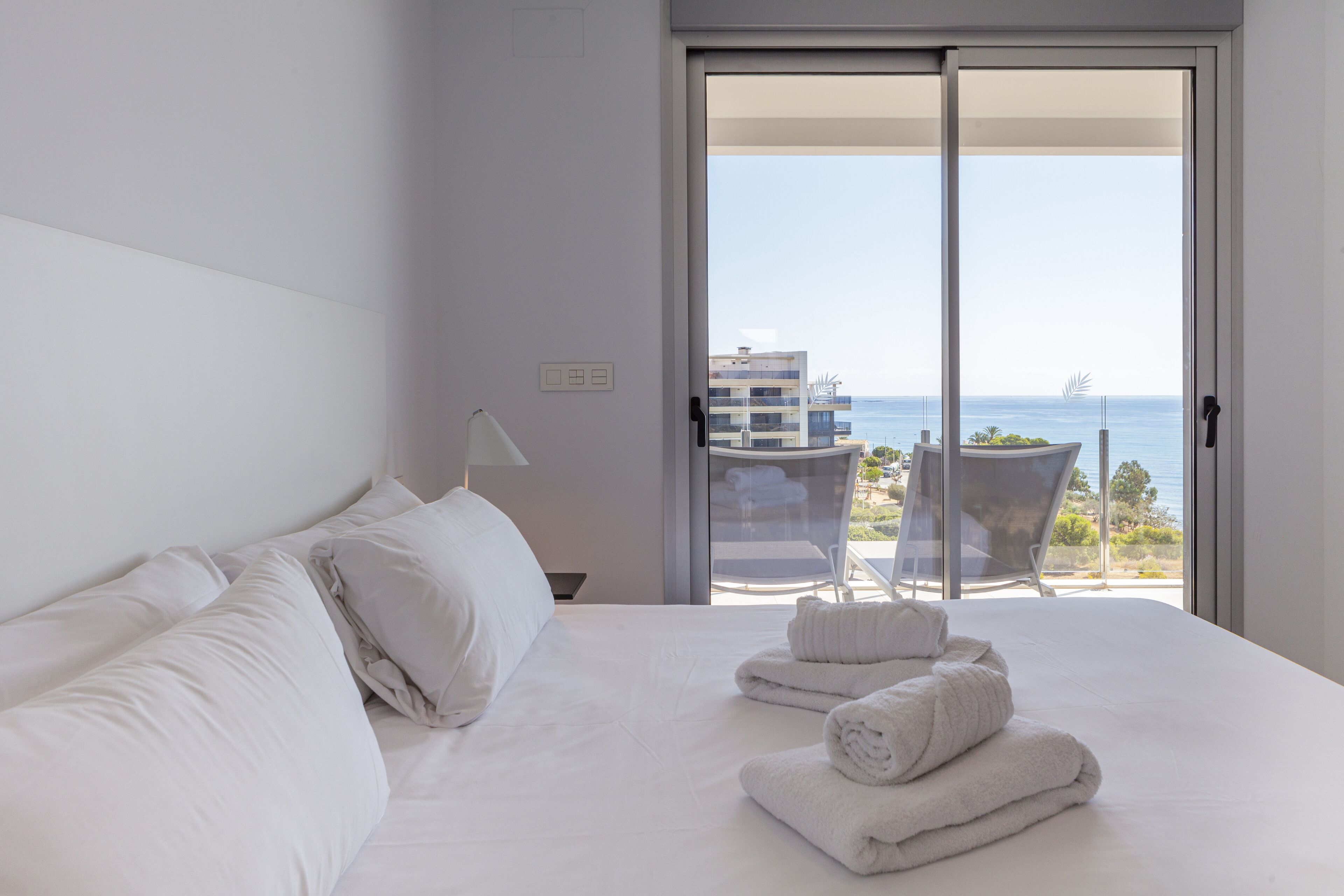 A hotel room with a bed, white linens, a lamp, and a view of the sea through a sliding door.