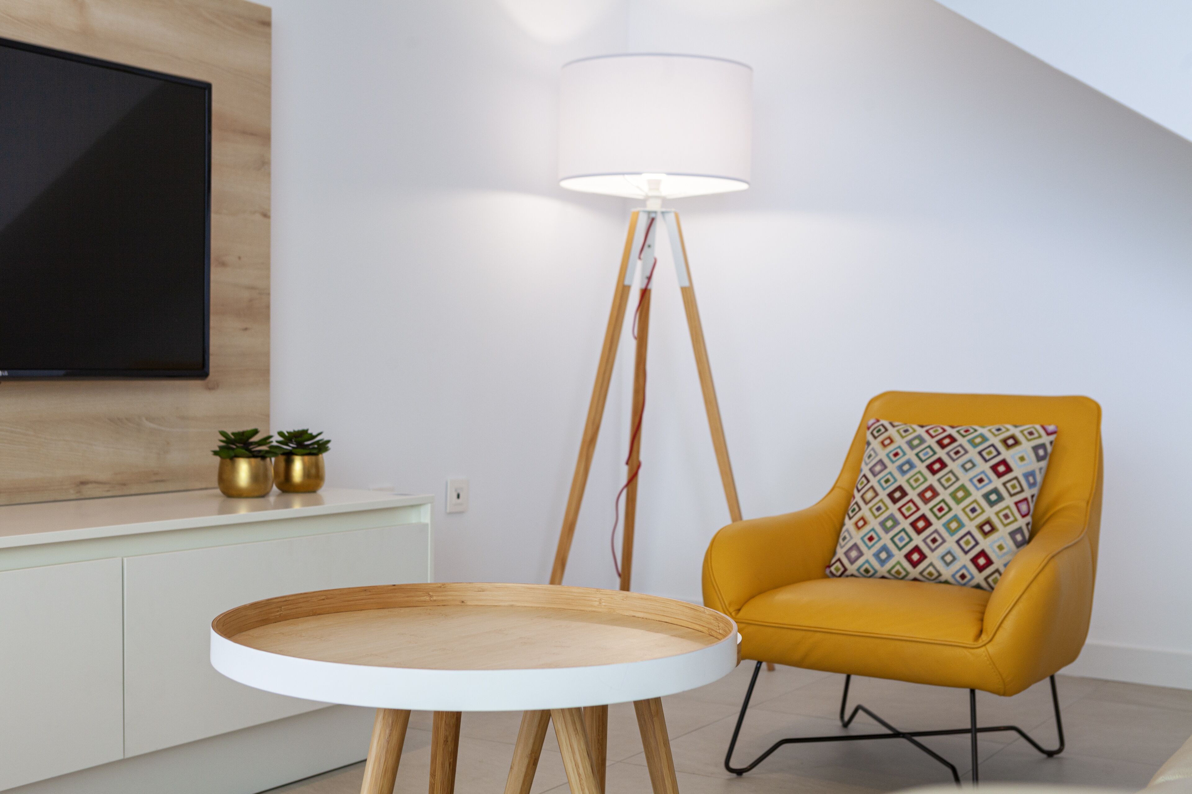 A modern living room with a round wooden table, a yellow armchair with a patterned cushion, a floor lamp, and a flat-screen TV mounted on the wall.