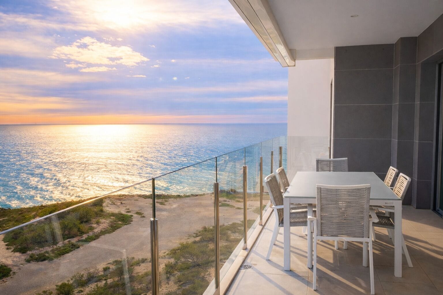 A balcony with a glass railing overlooking a beach and ocean, a table with chairs, and a modern interior design.