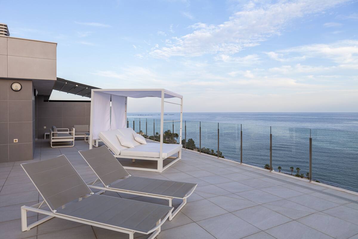 Eine Dachterrasse mit zwei Liegestühlen und einem Baldachinbett mit Blick aufs Meer.