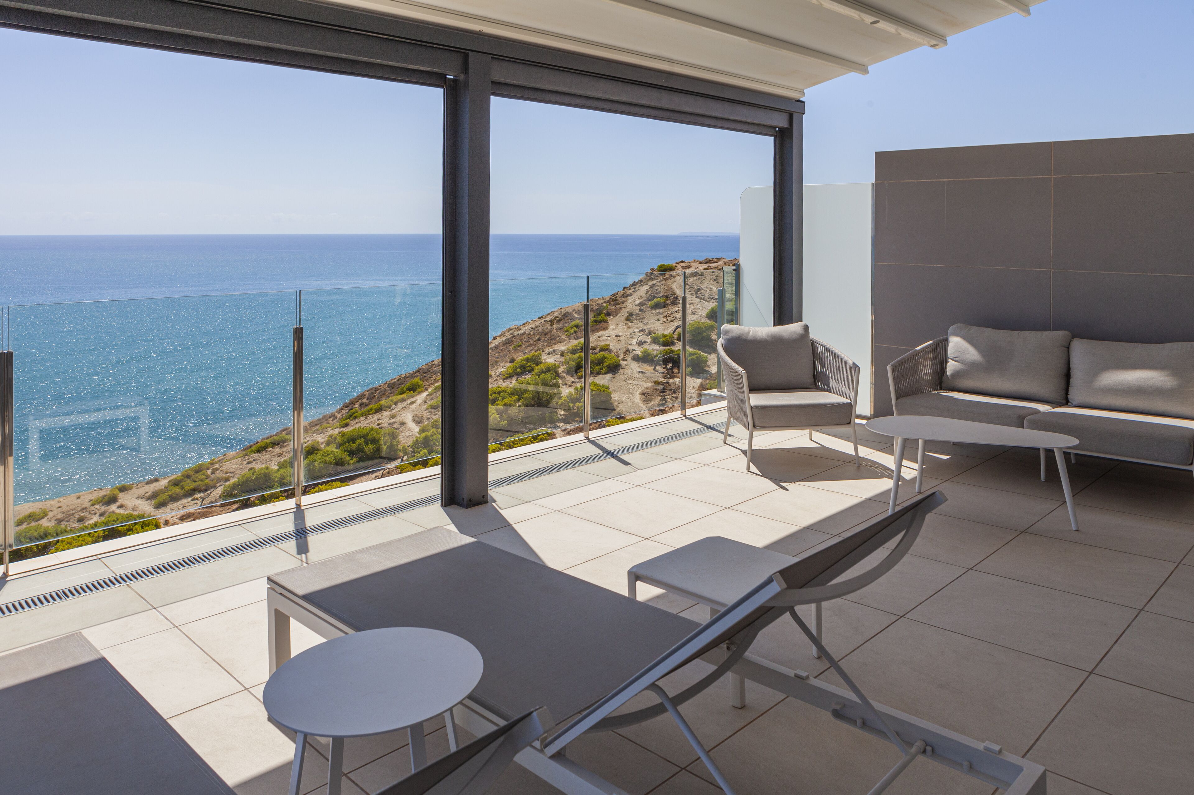 A modern outdoor balcony with a view of the sea, furnished with a sofa, chairs, and a table.