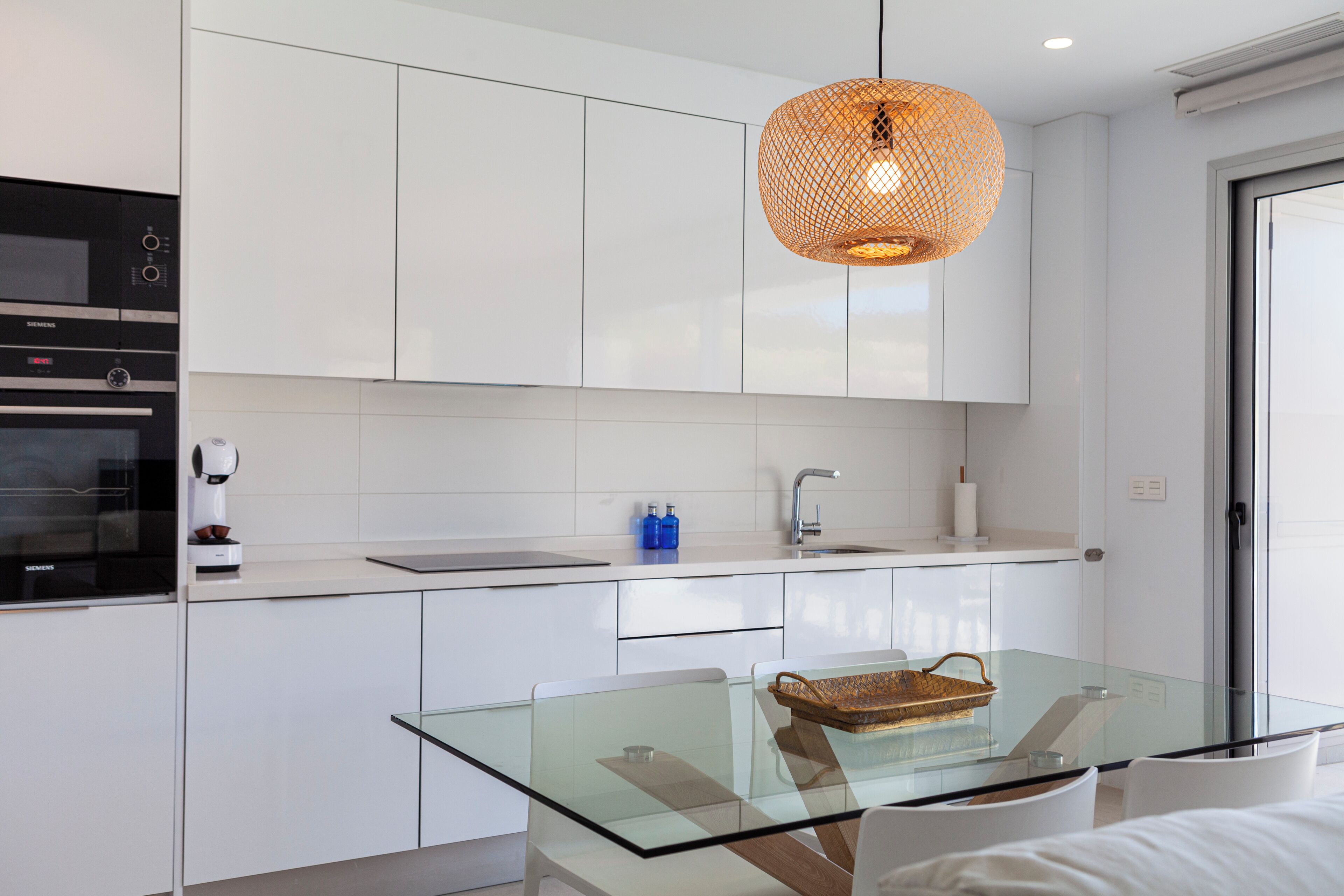 Cozinha moderna com armários brancos, mesa de jantar com tampo de vidro e luminária pendente.