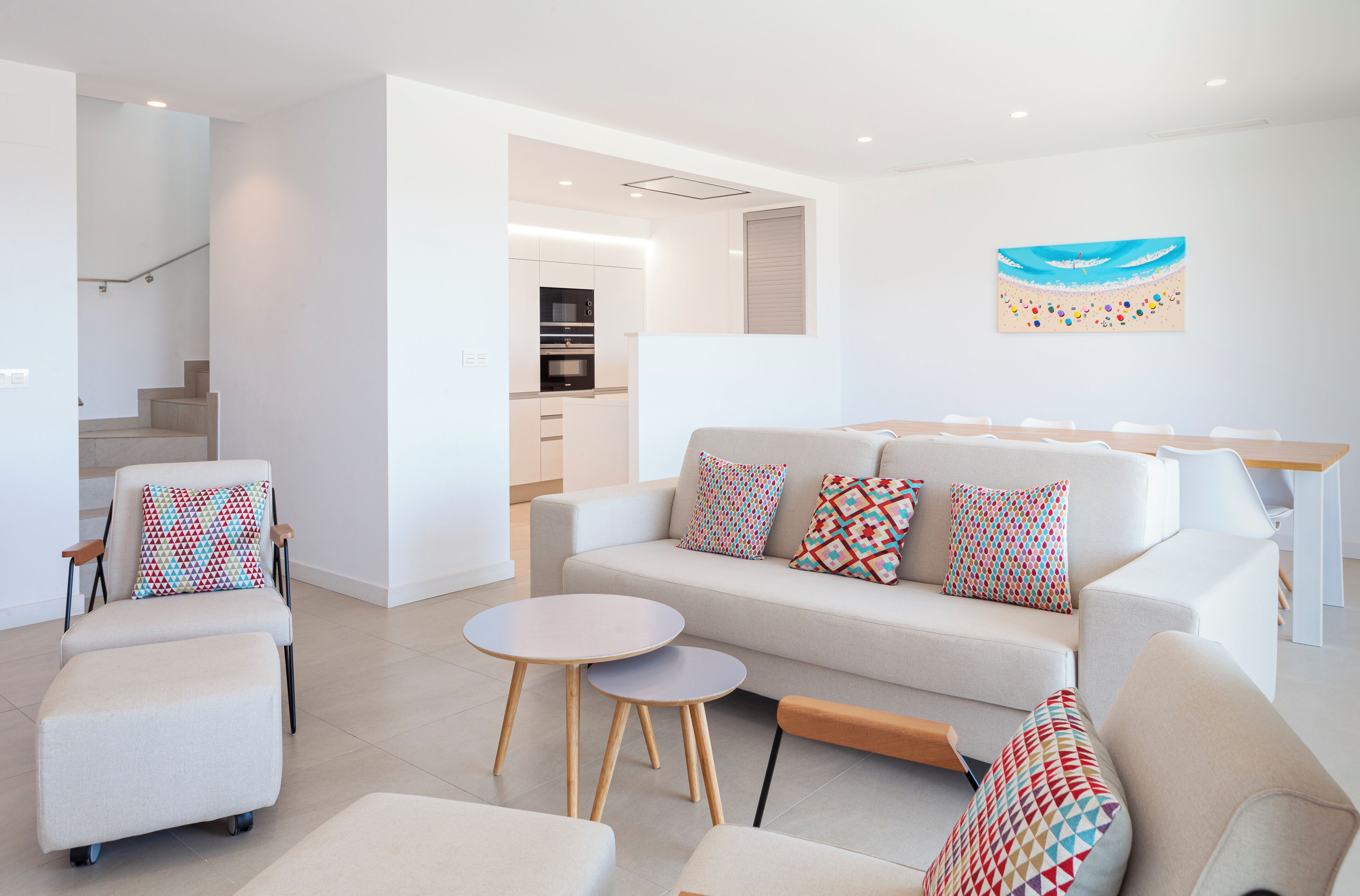 Un salon moderne avec un canapé, des fauteuils et une table basse. On aperçoit un escalier menant à un niveau supérieur, et une cuisine est visible au fond.