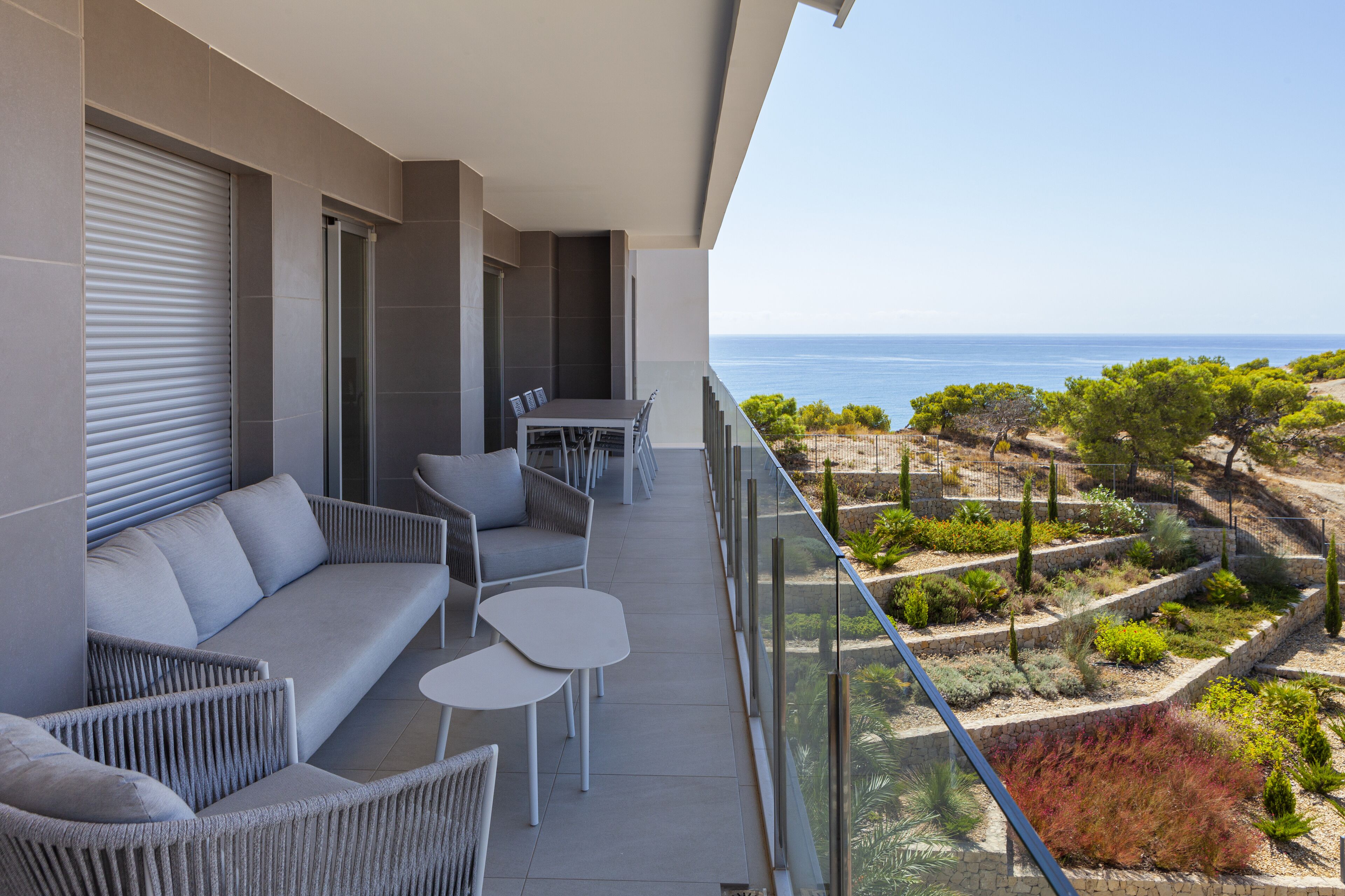 A balcony with a sofa, chairs, and a table, overlooking a landscaped garden and the sea.