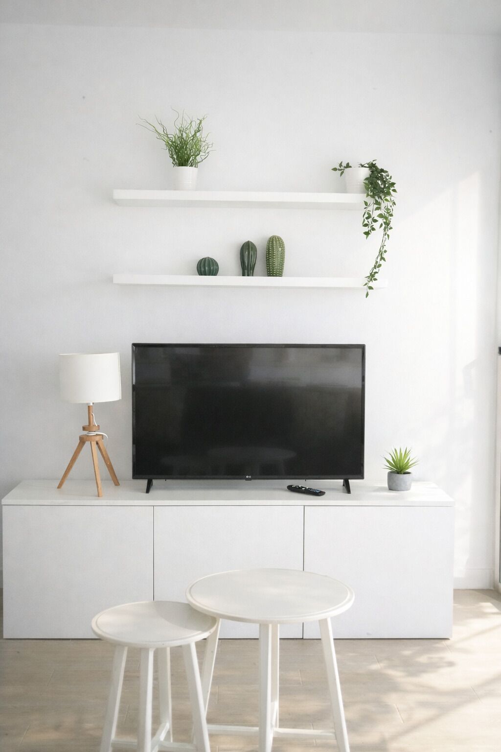 A modern living room with a flat-screen TV, white shelves with plants, a lamp, and a small table.