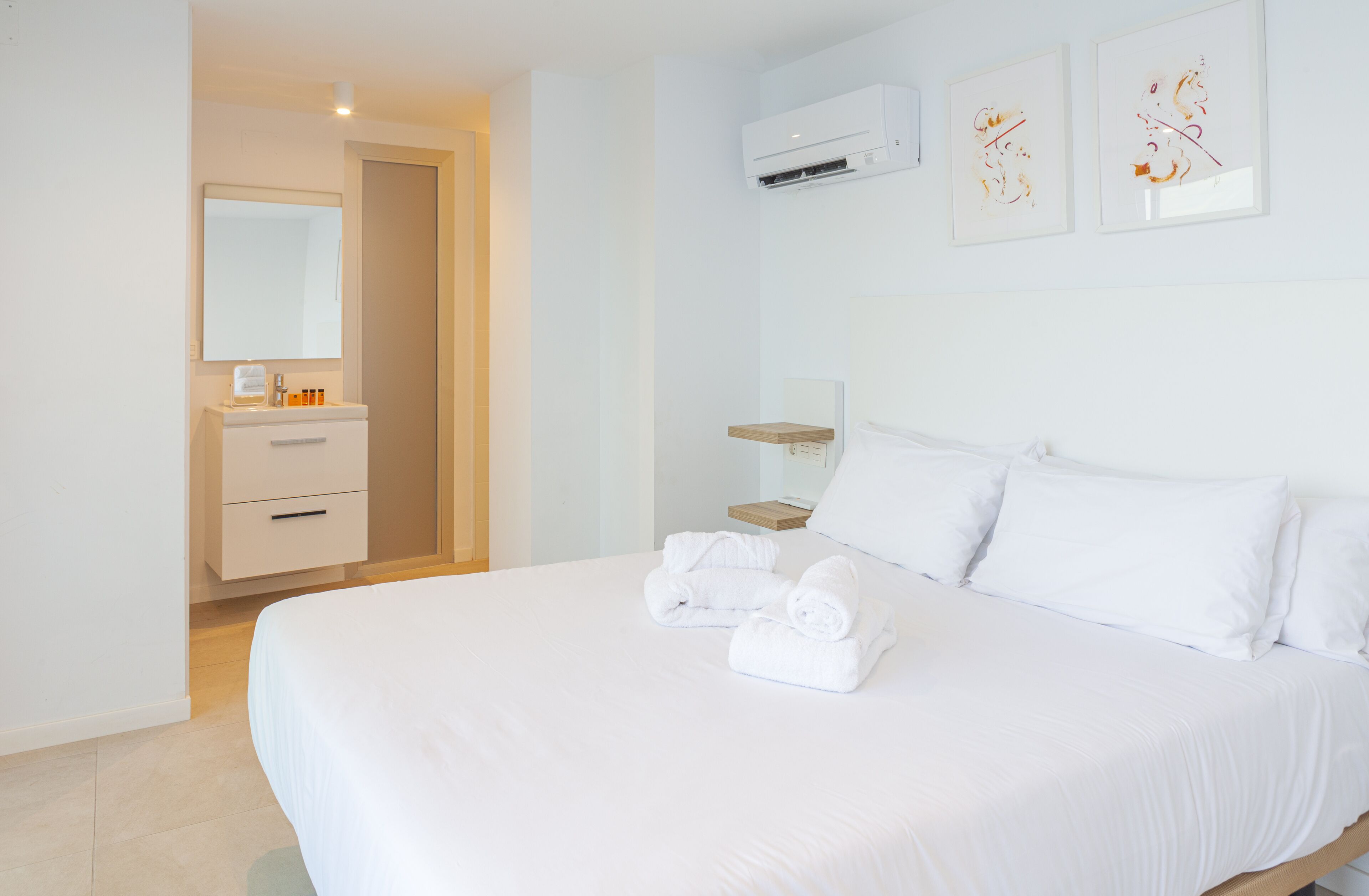 A hotel room with a large bed, a bedside table, a mirror, a wall-mounted air conditioner, and two framed artworks.