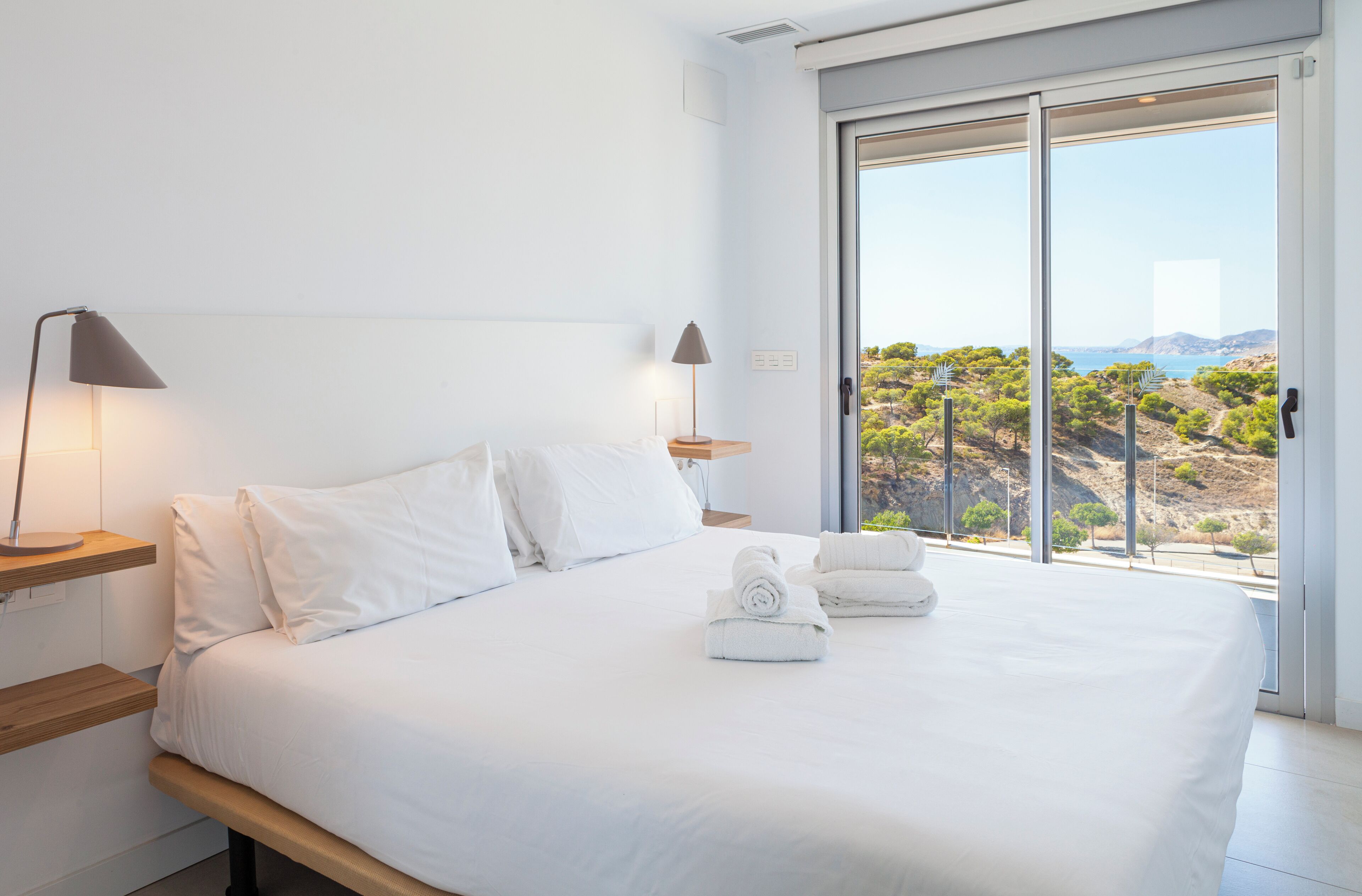 Una habitación de hotel moderna con una cama grande, dos mesitas de noche con lámparas y un balcón con vistas panorámicas.