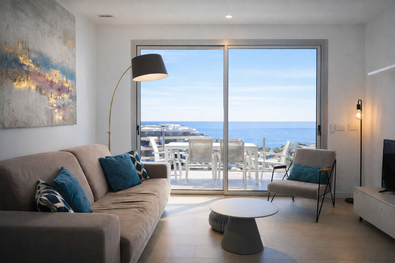A modern living room with a sofa, coffee table, and chairs, a large window with a view of the sea, and a painting on the wall.