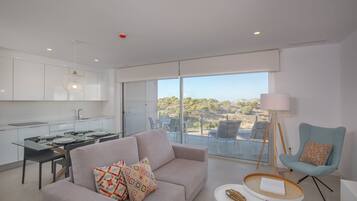 A modern living room with a sofa, a coffee table, and a chair. There is a dining table with chairs and a kitchen area. A large window offers a view of the outdoors.