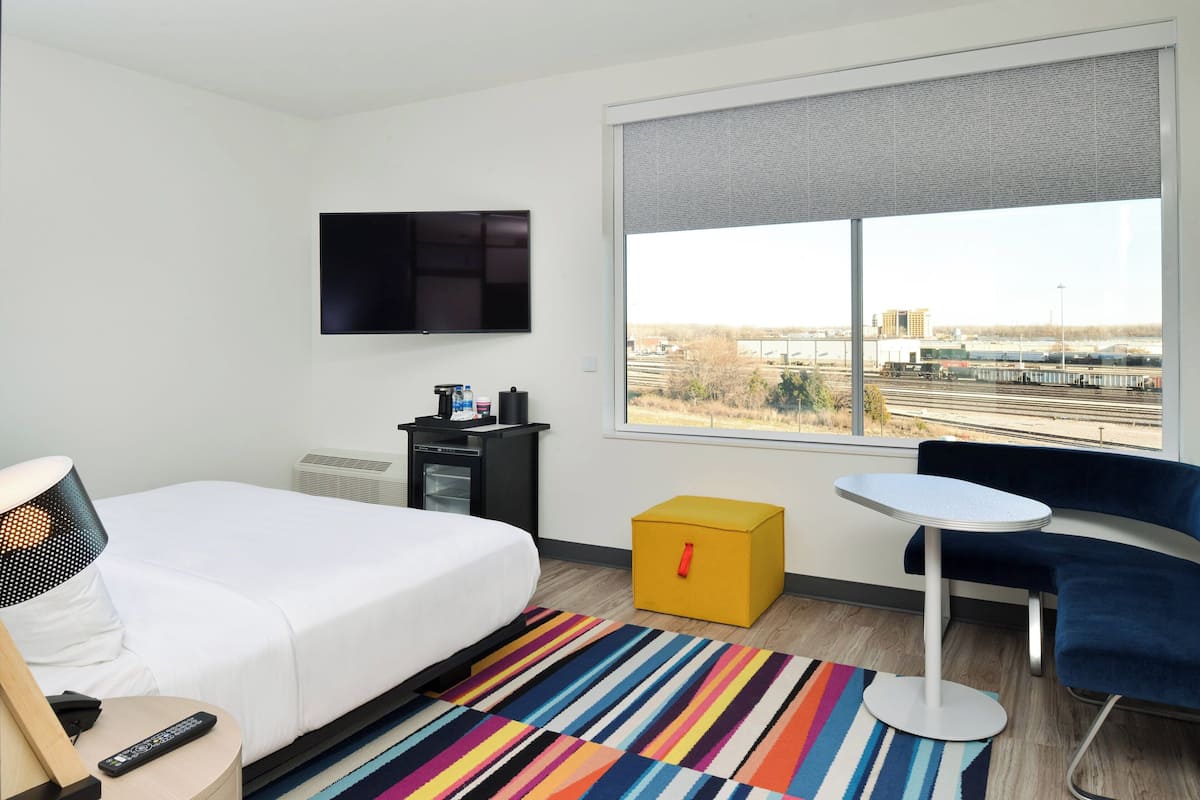 A hotel room with a bed, a TV mounted on the wall, a small table, a chair, and a large window with a view of a bridge and buildings.