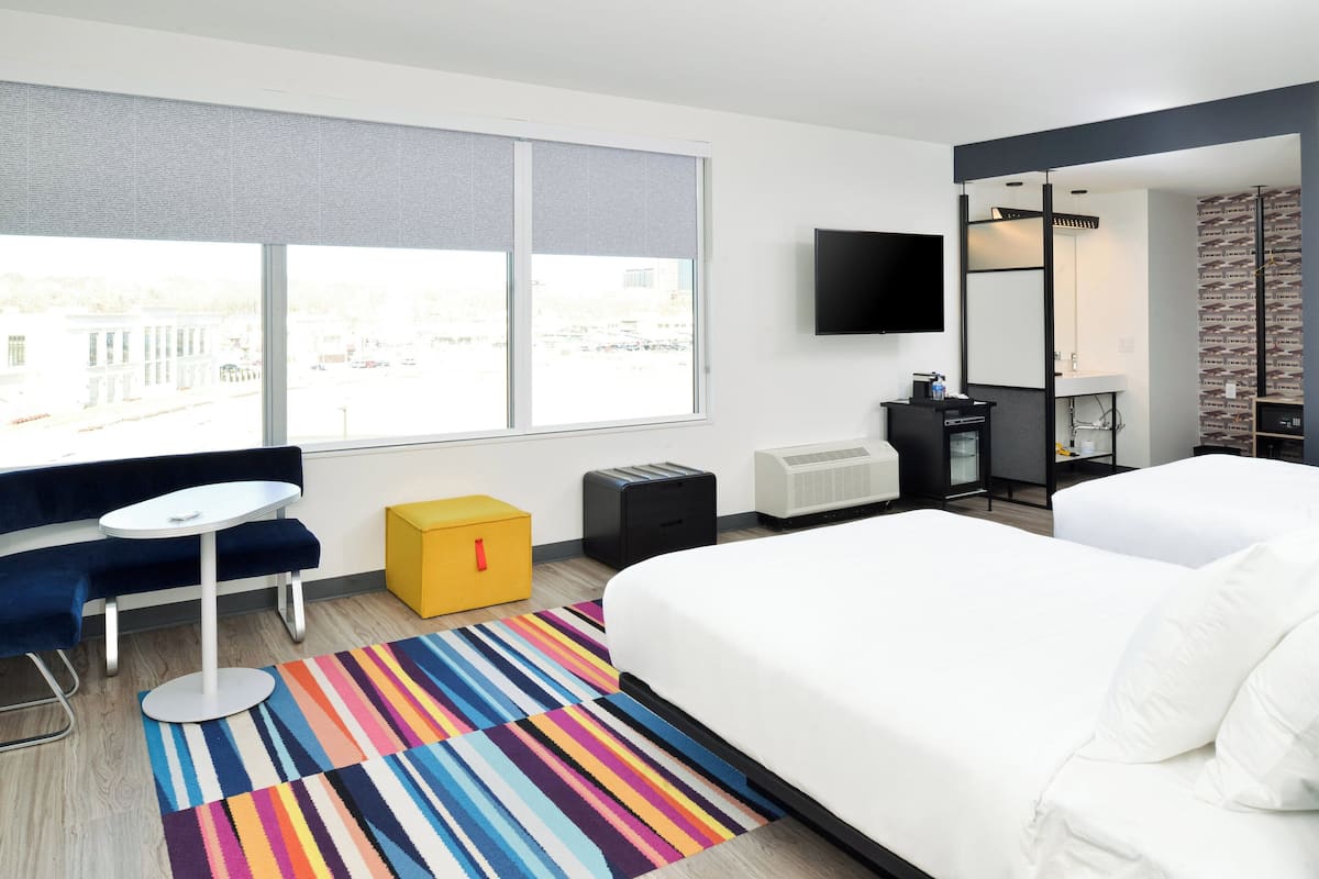 A hotel room with a large bed, a small table, a sofa, a colorful striped rug, a TV, and a window with blinds.