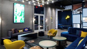 A modern lounge area with sofas, chairs, and tables, featuring a large illuminated sign and a glass door entrance. - Aloft by Marriott North Kansas City (North Kansas City)