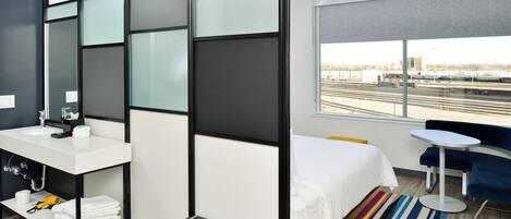 A modern hotel room with a white sink, a bed with white linens, a small table, a bench, and a large window with a view of train tracks.