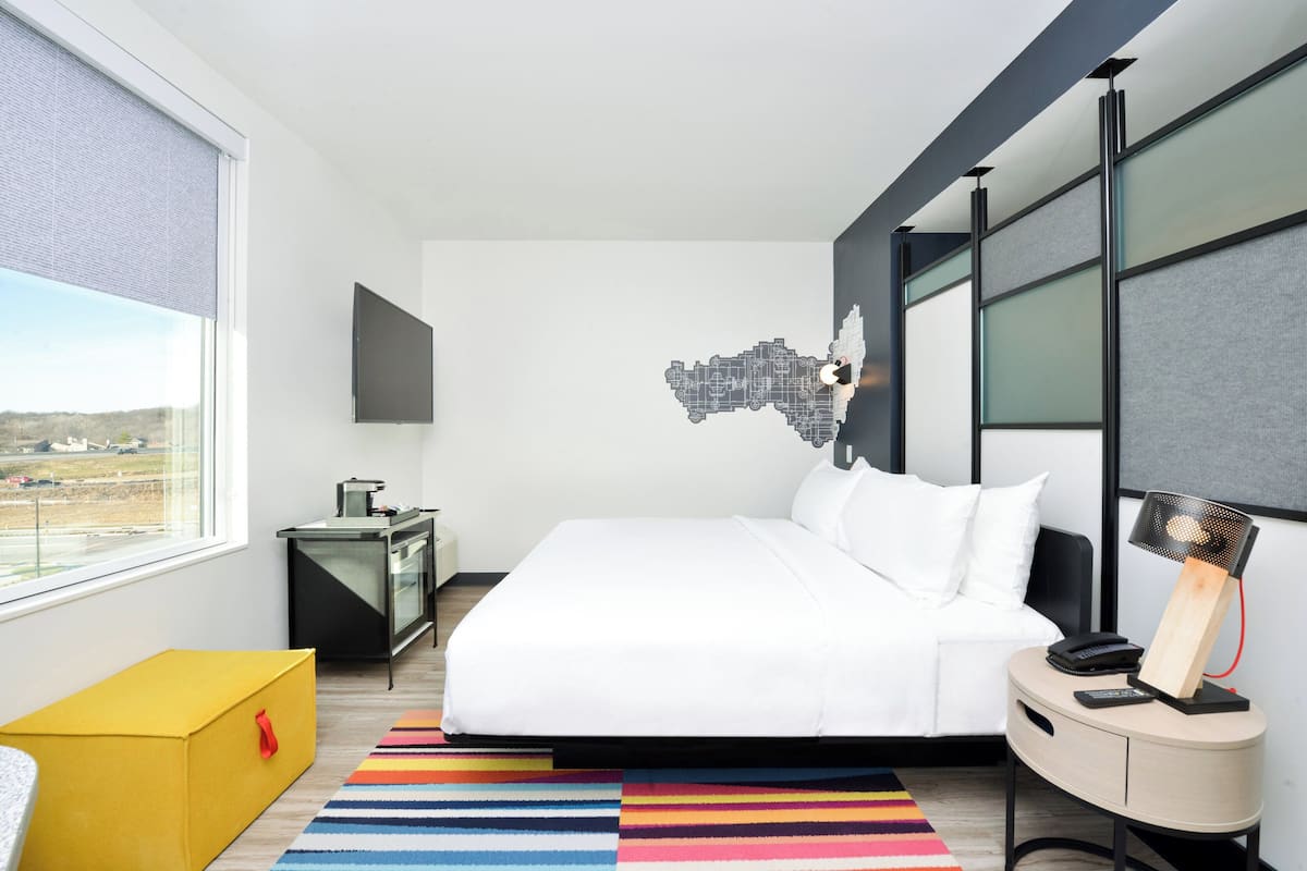 A modern hotel room with a large bed, a small desk, a chair, a colorful striped rug, and a cityscape wall art.