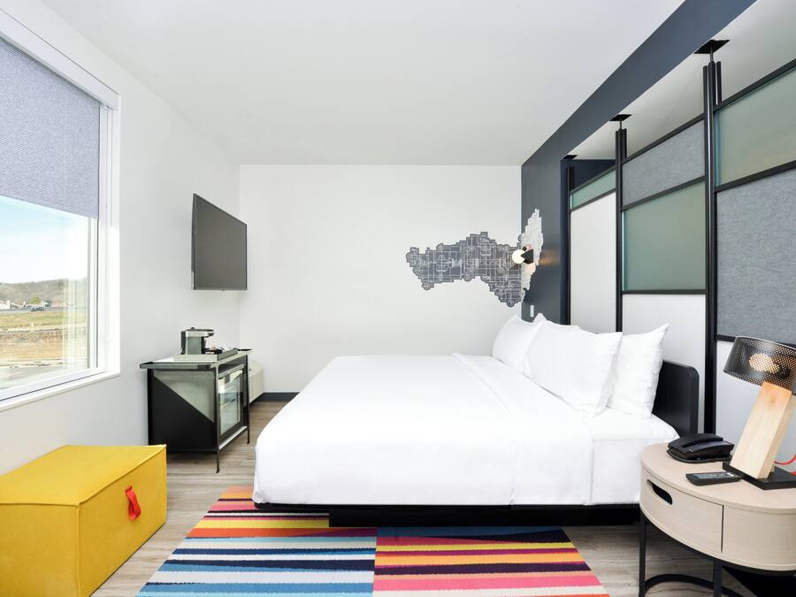 Una camera d'albergo moderna con un grande letto, una scrivania, una sedia, un tappeto colorato a righe e un'opera d'arte con una veduta sulla città.
