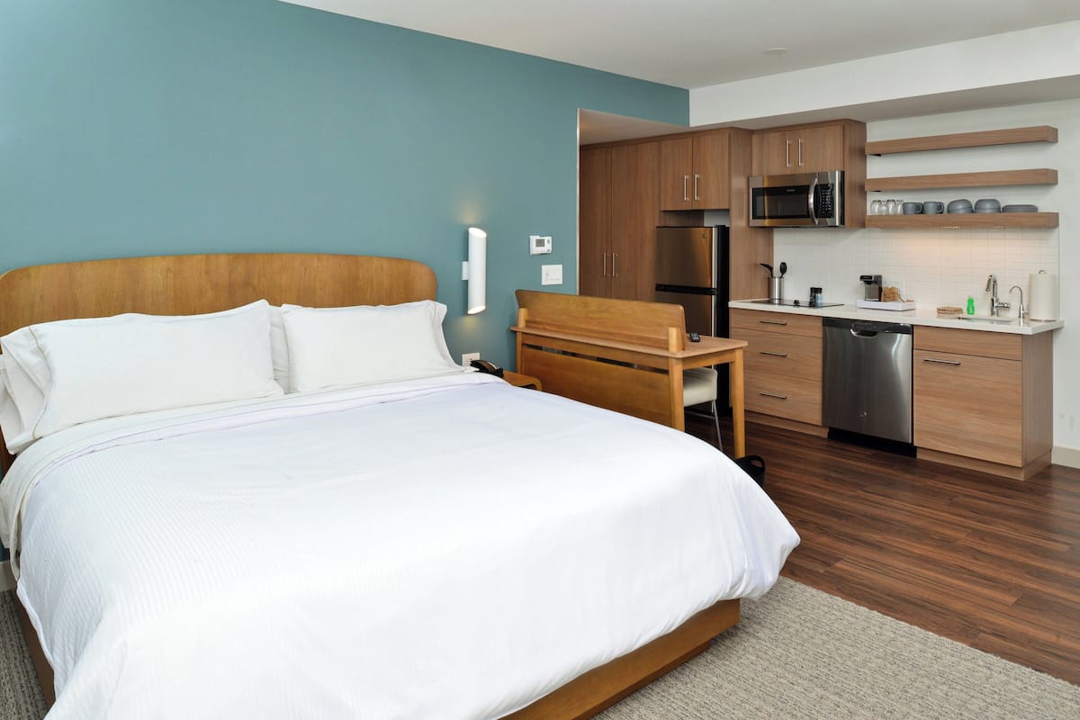 A modern bedroom with a large bed, wooden headboard, and a small desk with a chair. Adjacent is a kitchenette with a microwave, refrigerator, and a small sink.