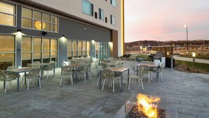 Un patio exterior con mesas y sillas, un hogar para fuego y un edificio con ventanas amplias al fondo.