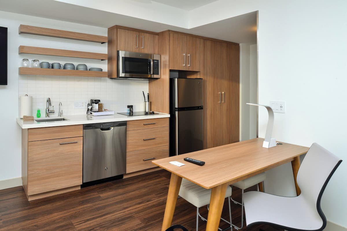 A modern kitchen with wooden cabinets, stainless steel appliances, a white countertop, and a dining area with a table and chair.
