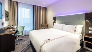 Habitación de hotel con una cama grande, un escritorio, una silla, un sillón pequeño y una mesita de noche con lámpara.