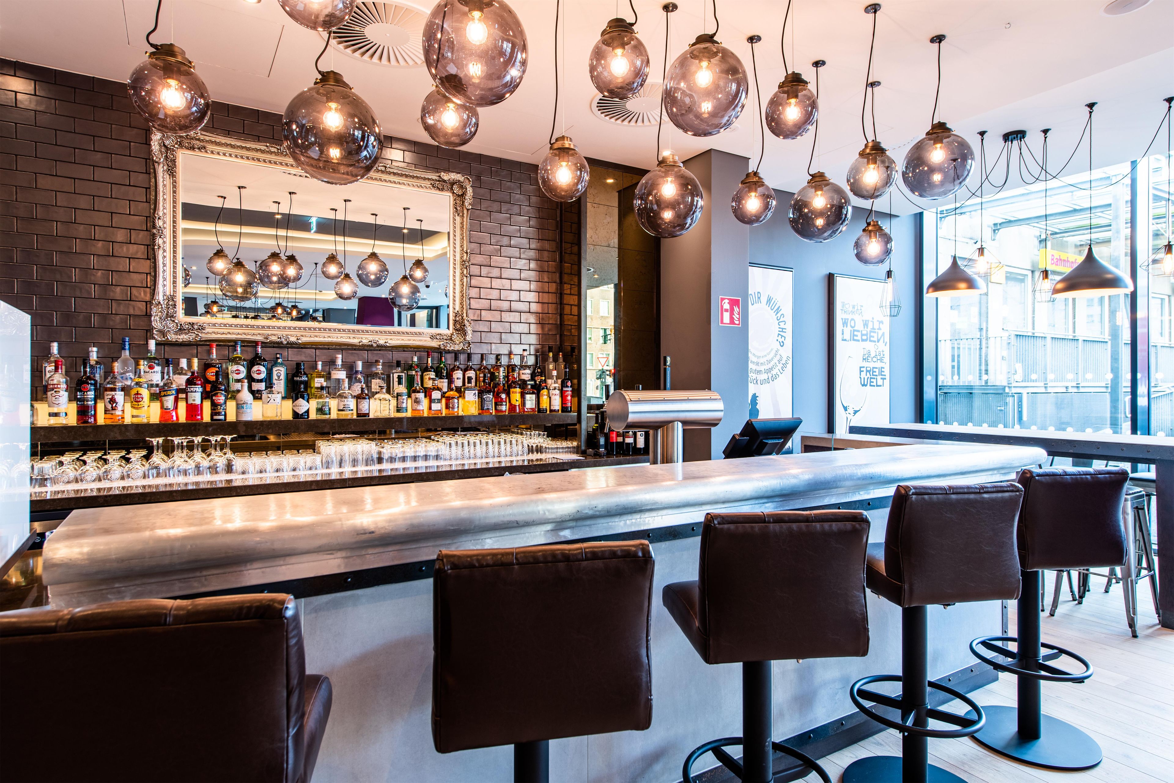 A bar area with a brick wall, a large mirror, hanging spherical light fixtures, and a bar counter with stools.