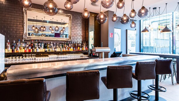 Zona de bar con pared de ladrillo, un espejo grande, lámparas esféricas colgantes y una barra con taburetes.
