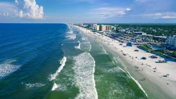 A coastal beach with a long stretch of sand, waves, and a row of buildings along the shore.