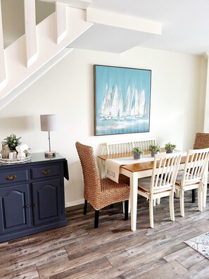 A dining area with a wooden table and chairs, a painting of sailboats on the wall, a lamp, and a cabinet with drawers.