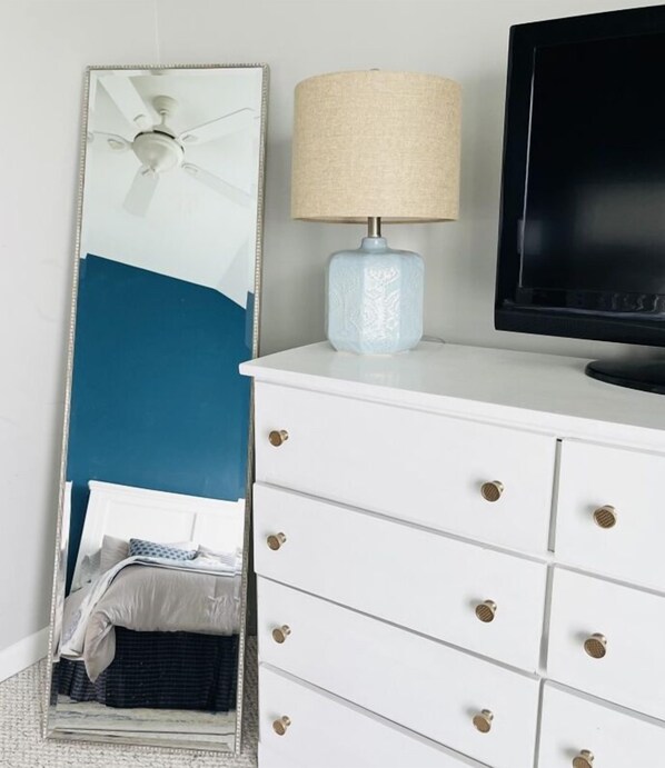 Une pièce avec une commode blanche, un miroir, une lampe et un téléviseur.