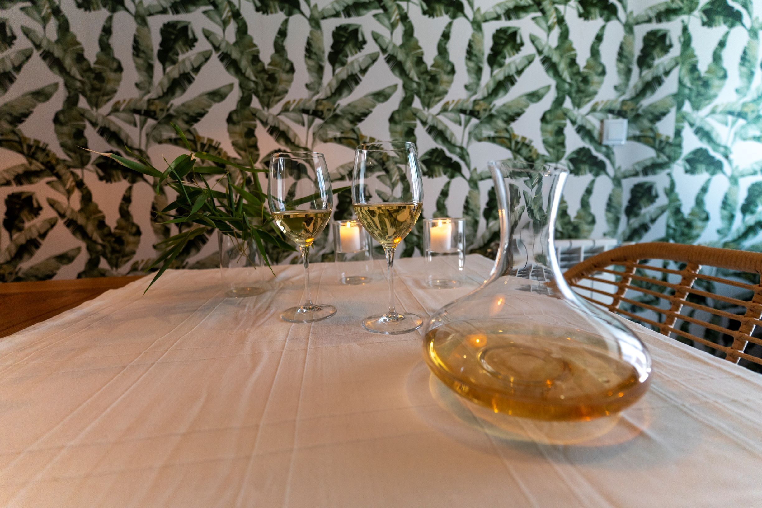 Una mesa servida con copas de vino y una botella, rodeada de velas, sobre un fondo de papel tapiz con hojas.