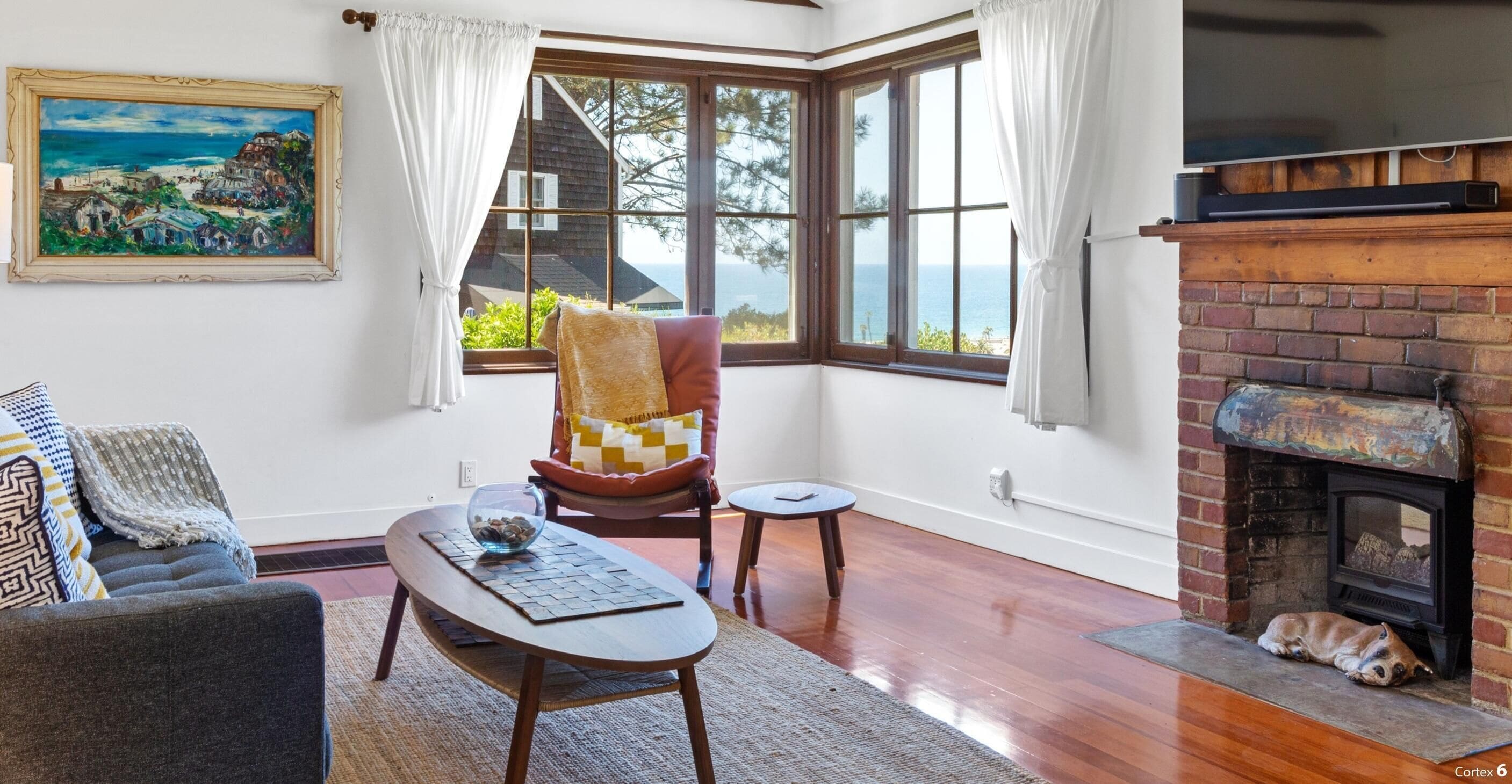 A cozy living room with a fireplace, a large window with a view of the sea, a comfortable armchair, and a sofa.