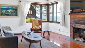 Ein gemütliches Wohnzimmer mit Kamin, einem großen Fenster mit Meerblick, einem bequemen Sessel und einer Couch.