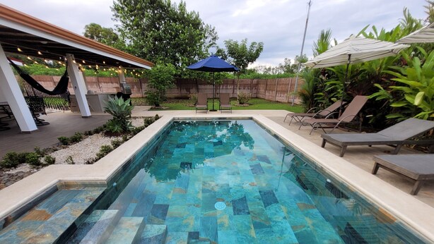 Una piscina rettangolare con decorazioni in piastrelle, circondata da un solarium in cemento. Si vedono anche sdraio e un gazebo con luci a tendina.