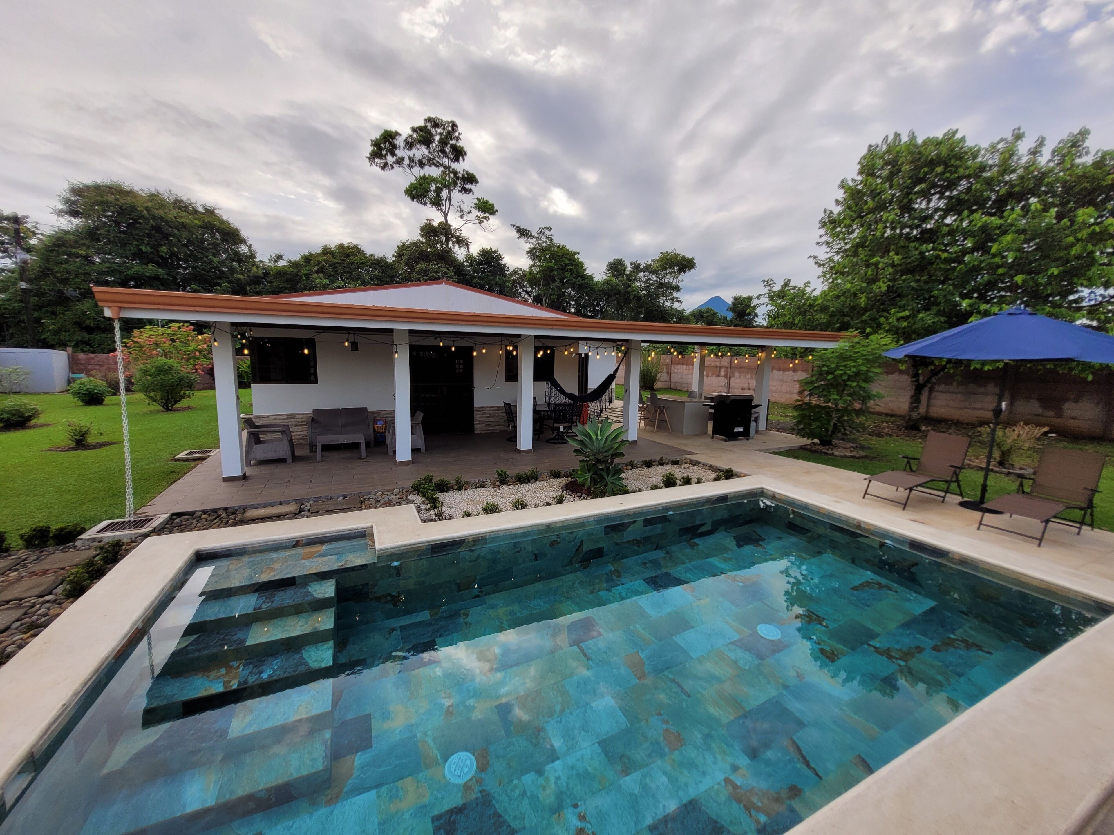 A backyard with a swimming pool, patio furniture, and a covered seating area.