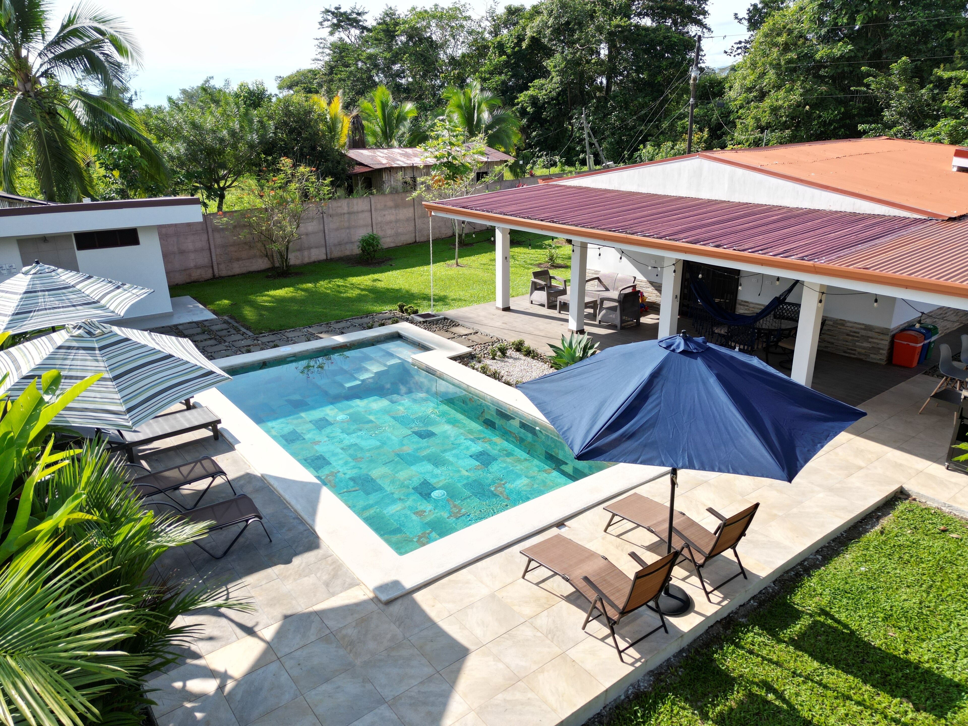 A swimming pool with a tiled deck, surrounded by lounge chairs and umbrellas, with a covered patio area and a view of a lush garden and trees.