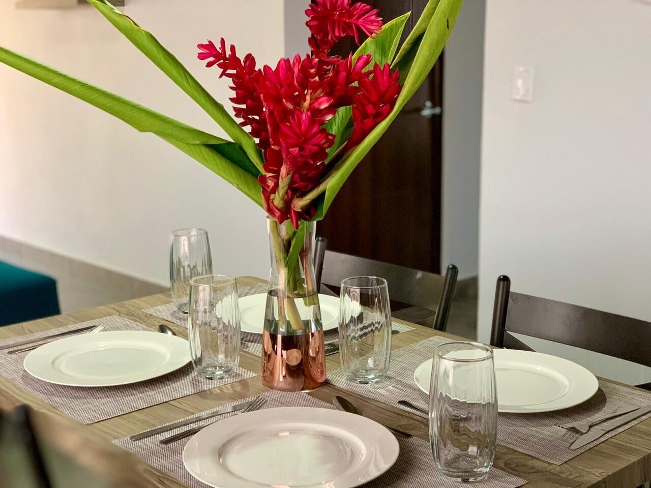 A dining table set with white plates, clear glasses, and a copper vase with red flowers.