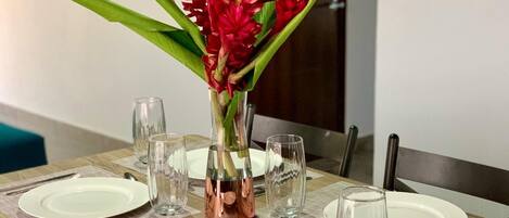 A dining table set with white plates, clear glasses, and a copper vase with red flowers.