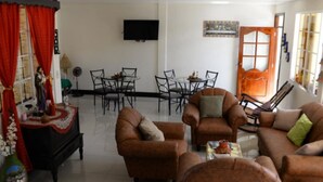 Sala de estar com sofás de couro marrom, uma mesa de jantar com cadeiras, televisão montada na parede e porta com painéis de vidro.