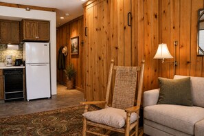A room with a wooden wall, a wicker chair, a sofa, a lamp, and a kitchen area with a refrigerator and coffee maker.