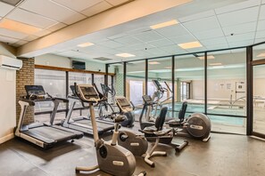 Ein Fitnessstudio mit Laufbändern, Crosstrainern und einem durch Glastüren einsehbaren Poolbereich.