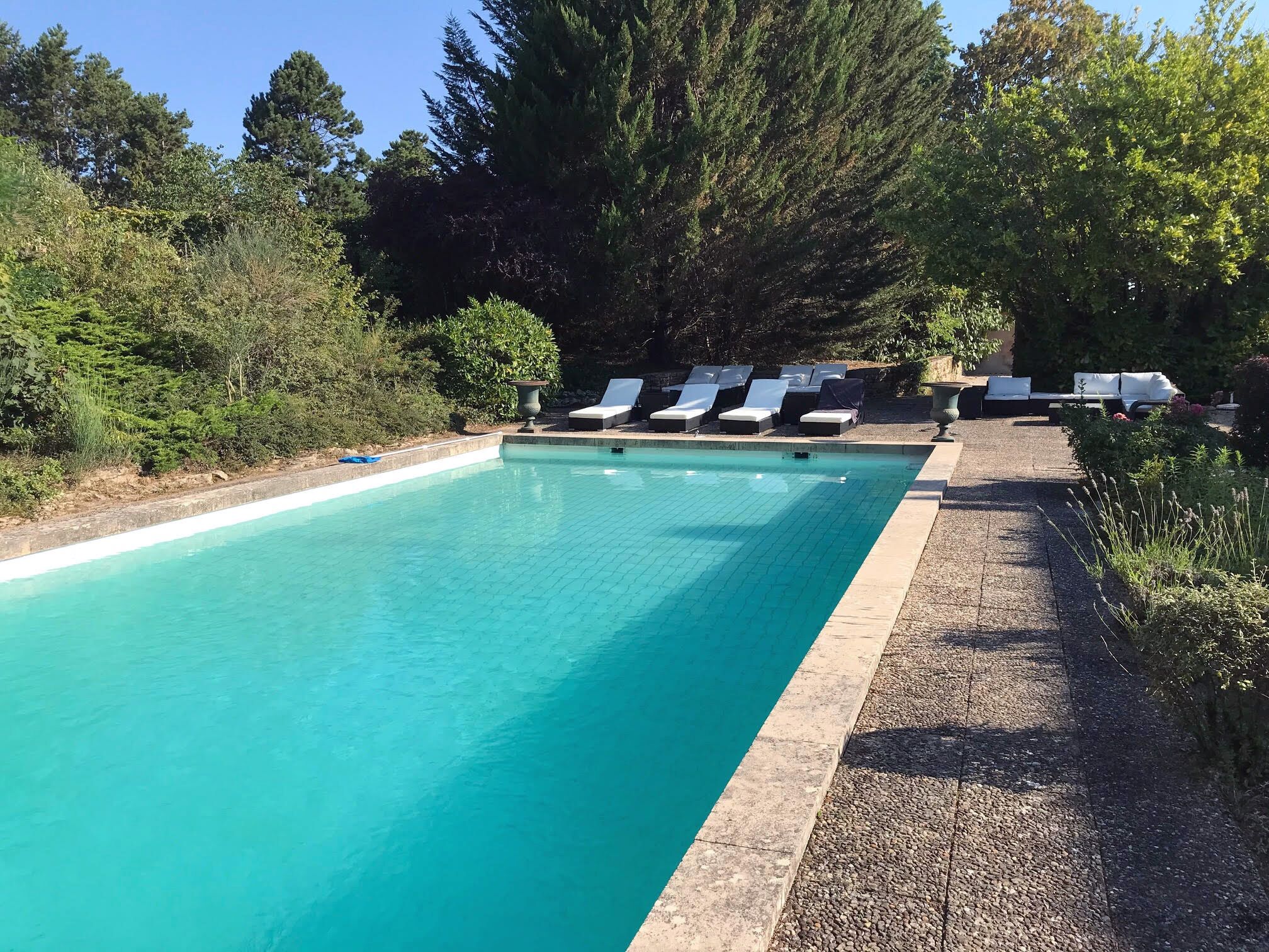 Une piscine rectangulaire à l’eau d’un bleu limpide, entourée d’une terrasse bordée de galets. Plusieurs chaises longues sont alignées le long du bord de la piscine.