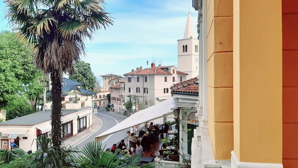 A street view with buildings, a palm tree, and a café with outdoor seating. - 50m vom Meer entfernt (Lovran)